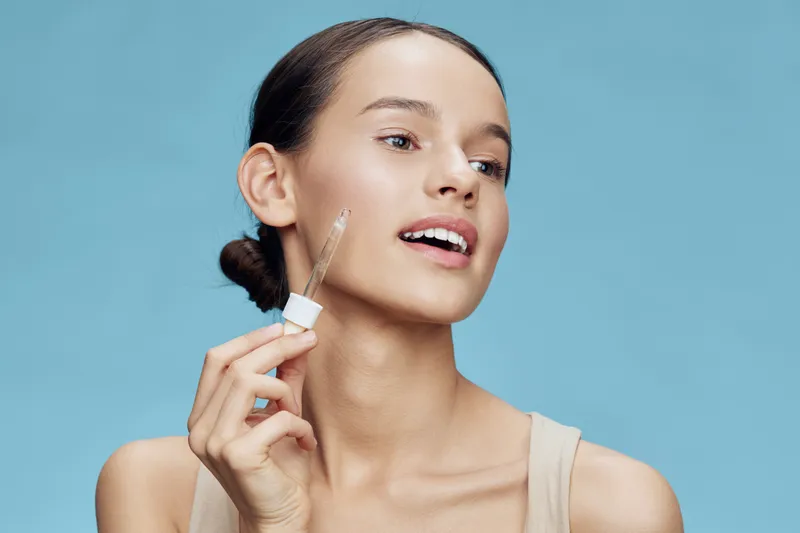 Young woman applying skincare serum on her face, showcasing glowing skin and a fresh look with soft blue background Perfect for beauty and wellness concepts