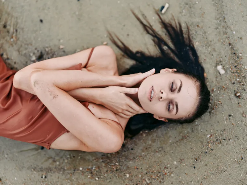 A woman lies on sandy ground with eyes closed, hands near her neck, and a serene expression, captured in warm light highlighting a calm, peaceful moment.