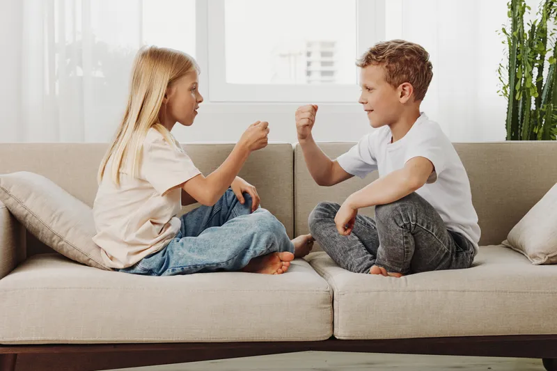 Two children sitting on a beige sofa play rock paper scissors game with focused expressions in a bright living room. Concept for childhood friendship, playtime, and family bonding moments.