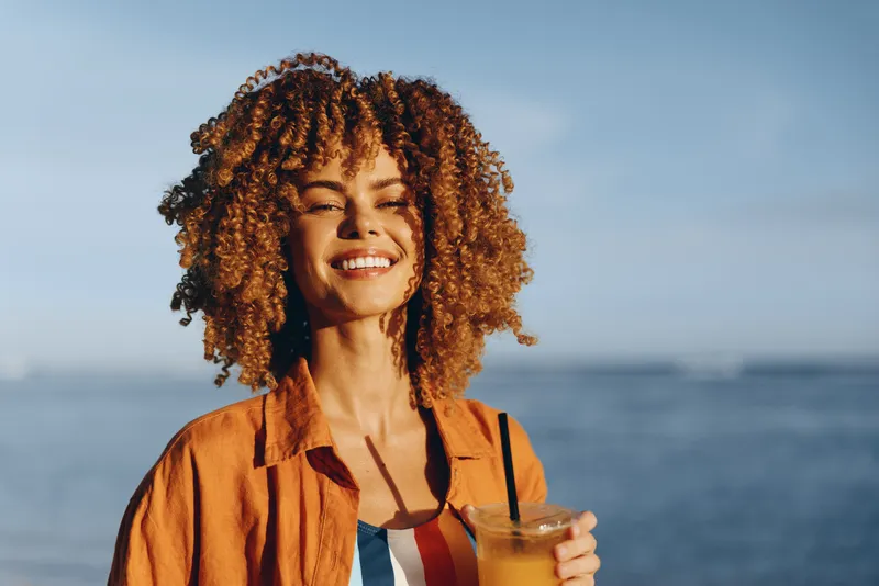 Curly hair woman smiling, holding orange drink cup, warm sunlight at seaside, outdoors with ocean and blue sky, casual vacation lifestyle, relaxed calm mood, close portrait