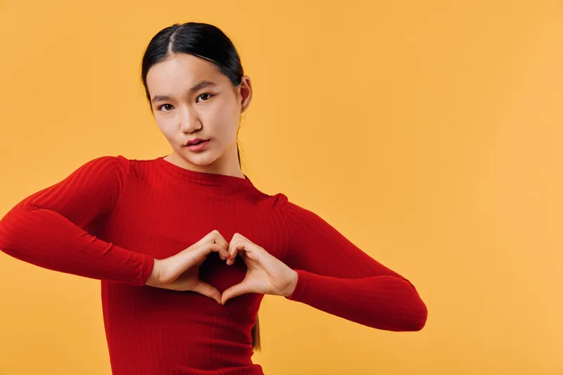 Woman in red sweater making heart shape with hands on orange background, expressing love and positive emotion with confident facial expression, studio portrait.