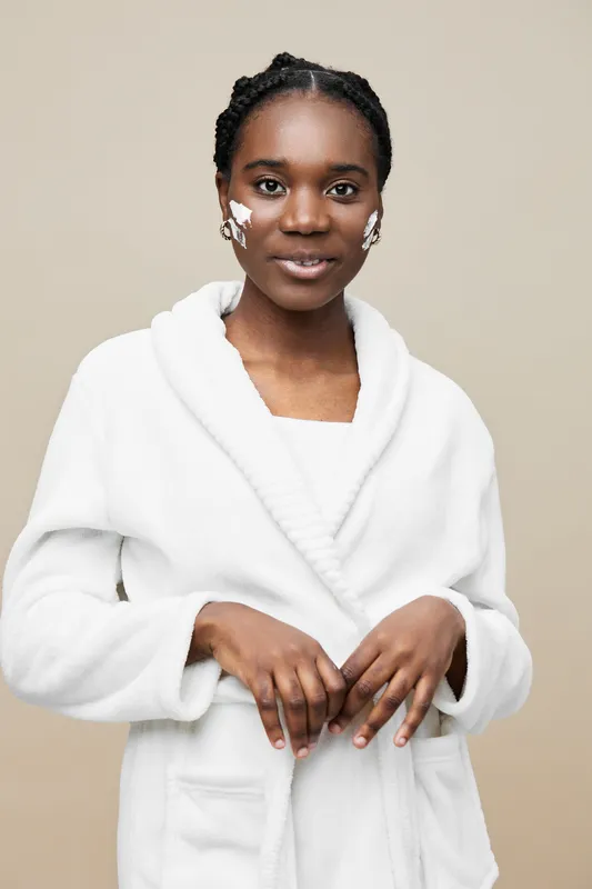 Young Black woman in a white robe applying skin care product, smiling against a neutral beige background, representing beauty and self care