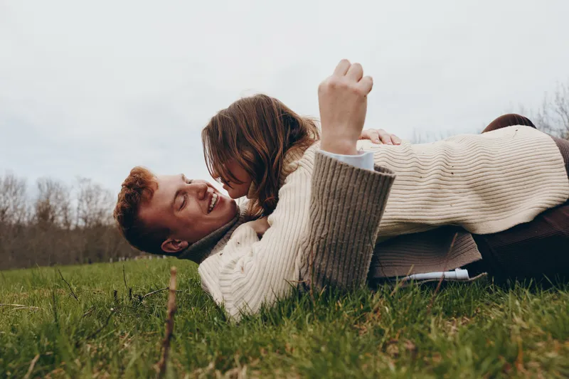 A couple outdoors, laughing and embracing on a grassy meadow, cozy sweater, natural colors, relaxed pose, candid moment, gentle breeze, warm connection, sunlit field in soft morning light.