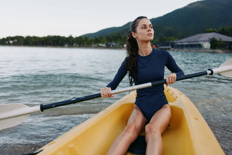 Athletic woman paddles a yellow kayak on calm water, wearing a dark blue swimsuit, poised with an oar as distant mountains rise and a soft cloudy sky frames the scene.