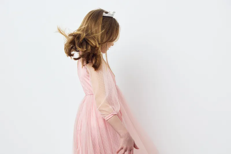 Playful young girl in a pink dress twirling, joyfully smiling, against a soft white background, evoking innocence and happiness in childhood moments