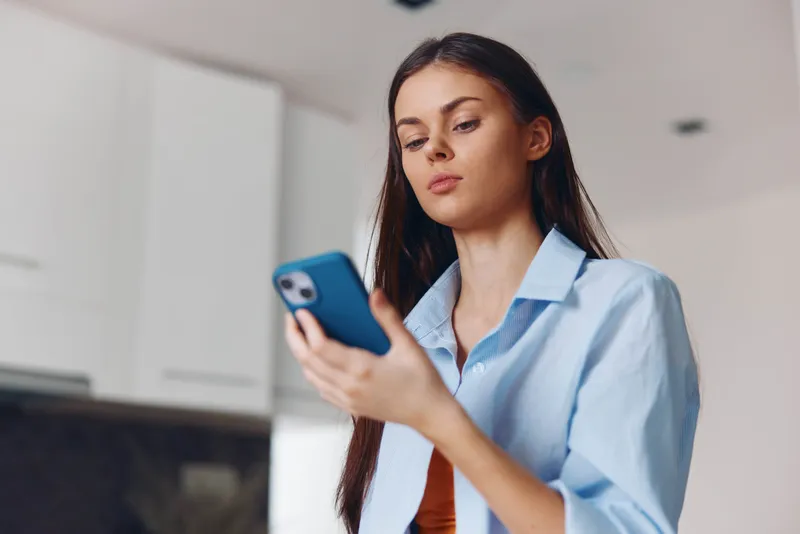 Женщина использует смартфон на кухне дома, просматривая интернет и проверяя сообщения