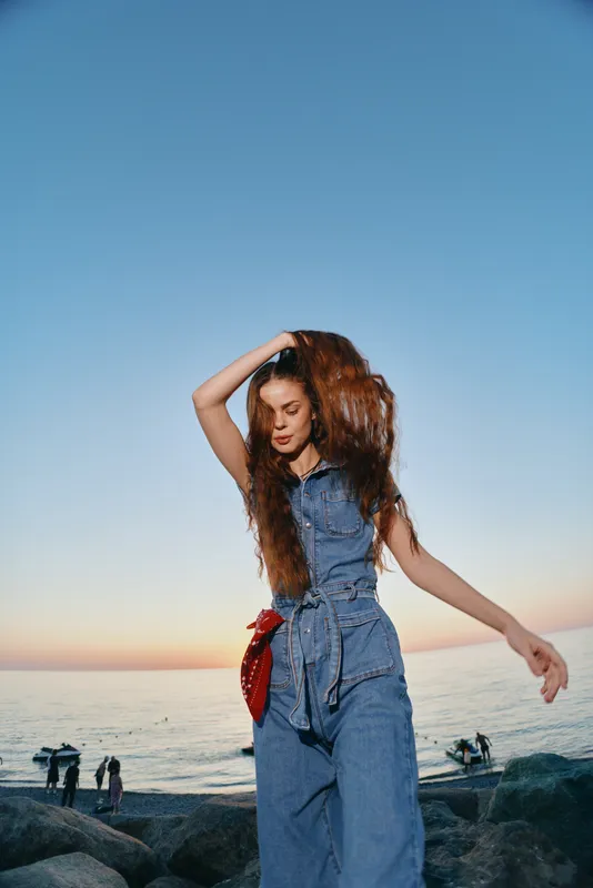 Lifestyle woman in modern boho-western denim street style dances freely by the ocean at sunset, showcasing film color tones and relaxed summer vibes in casual outfit.