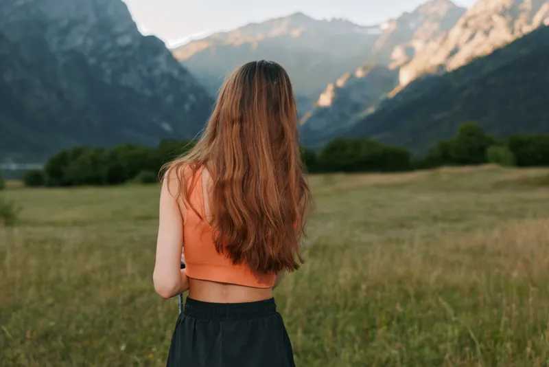 A young woman stands in a wide open field facing distant mountains. She wears an orange crop top and dark shorts, sunlight softly illuminating the calm outdoor scene.