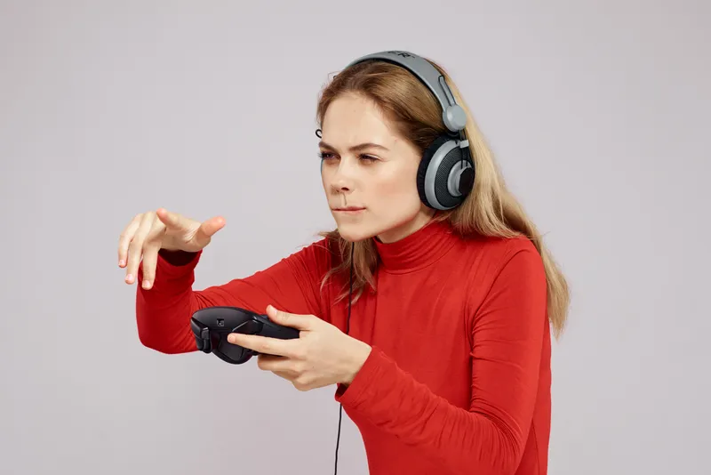 Женщина в наушниках с джойстиком в руках, играет в игры, весёлое времяпрепровождение, образ жизни, развлечения, красная рубашка, серый фон