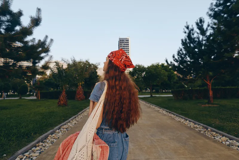 young woman with long red hair walking outdoor park wearing a red bandana and carrying tote bags on a clear day with green trees and modern buildings