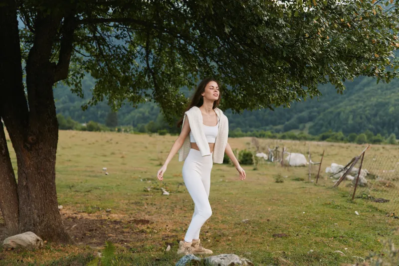 Woman in white walks through a sunny field beneath a large tree, enjoying a carefree outdoor lifestyle and a candid moment of nature and freedom