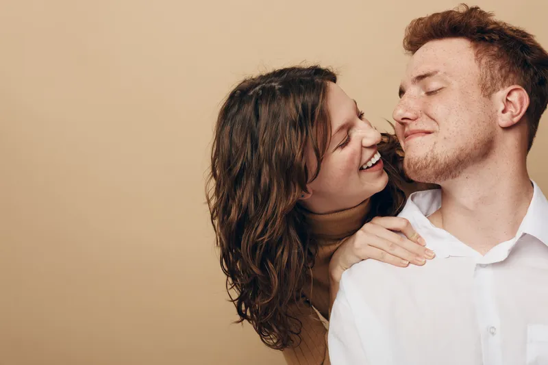 Happy young couple embraces and smiles with closed eyes against beige background. Concept of love, affection, relationship, and intimacy between man and woman.