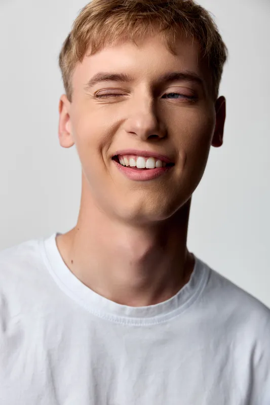 Closeup of a smile, wink youth in a casual white shirt, showing playful charm, relaxed mood and bright studio light for lifestyle, youth fashion and friendly expression