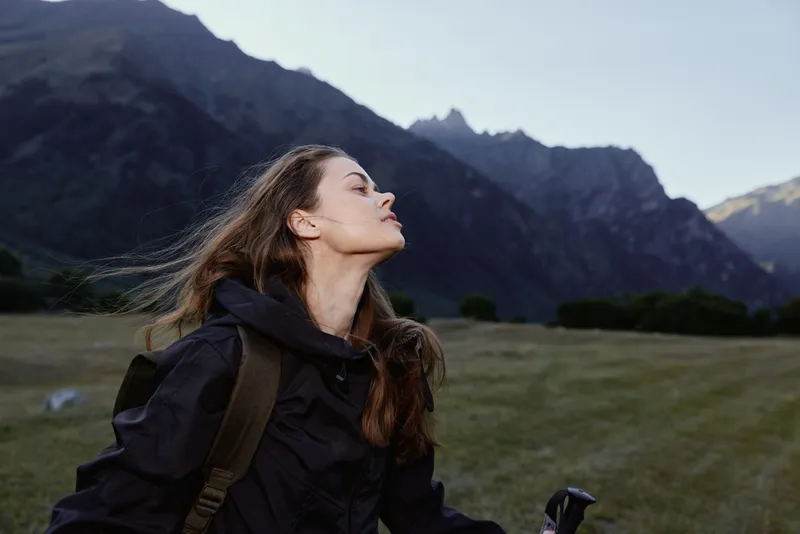 A woman outdoors, gazing upward with wind in her hair, wearing a dark jacket and backpack, framed by rugged mountains and an open sky, conveying exploration and serenity.