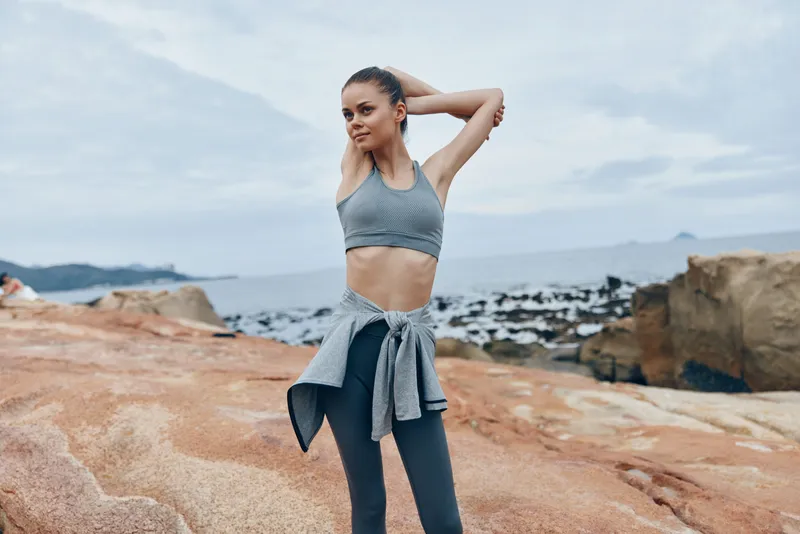 Young fit woman in workout gear stretching on rocky beach, showcasing athleticism and focus, with a calm sea backdrop and a gray color palette