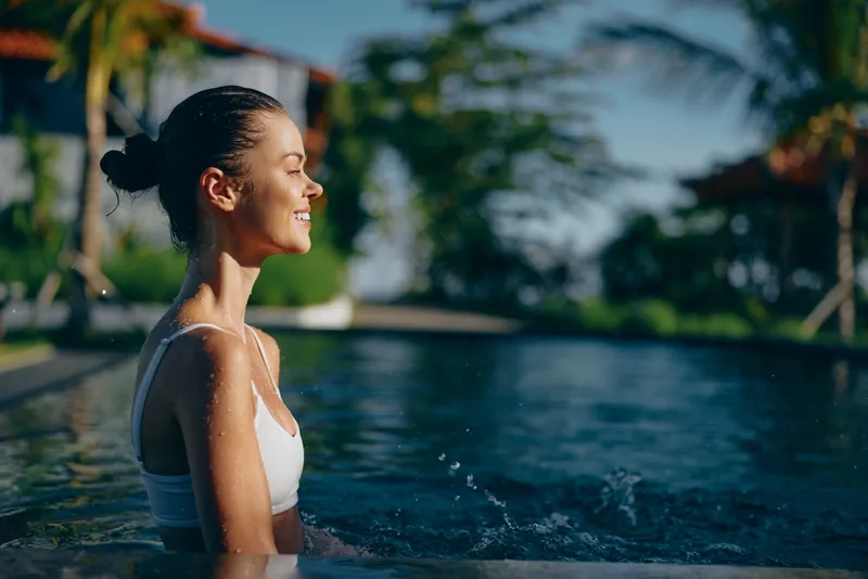 Joyful woman in a poolside moment, profile view, sunlit water and soft breeze, casual swimwear, relaxed posture, lush backdrop, warm tones, lifestyle vibe, candid outdoor scene.