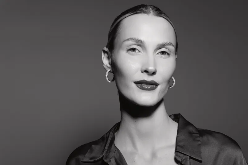 Portrait of a fair skinned Caucasian woman with slicked back hair and hoop earrings in black silk blouse on grey background.
