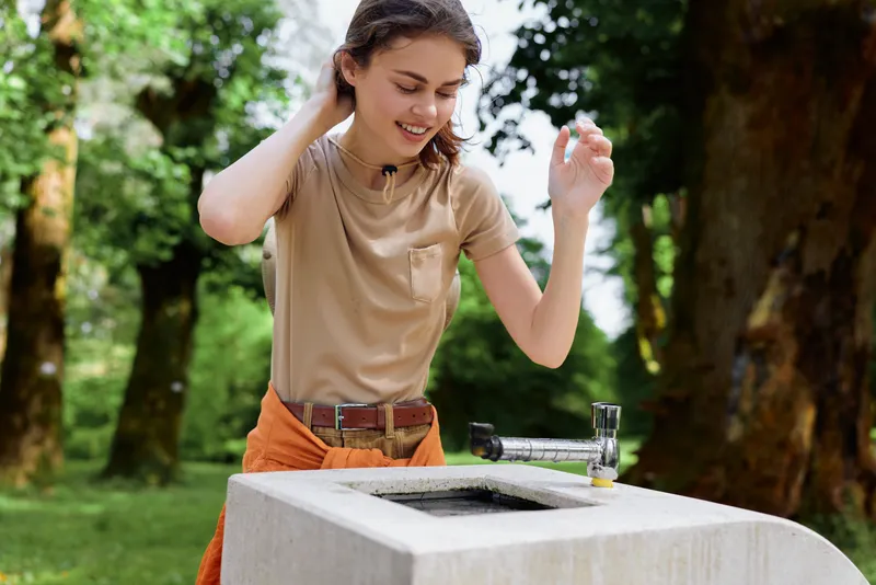 Woman outdoor park fountain with glass and tap, smiling while filling water at a stone trough. Casual clothes, green trees in background, natural summer hydration moment.
