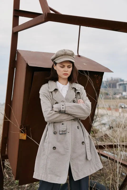 Young woman in checkered coat and matching hat stands with arms crossed outdoors near rusty metal structure. Overcast sky and dry grass create moody urban atmosphere and fashion concept.