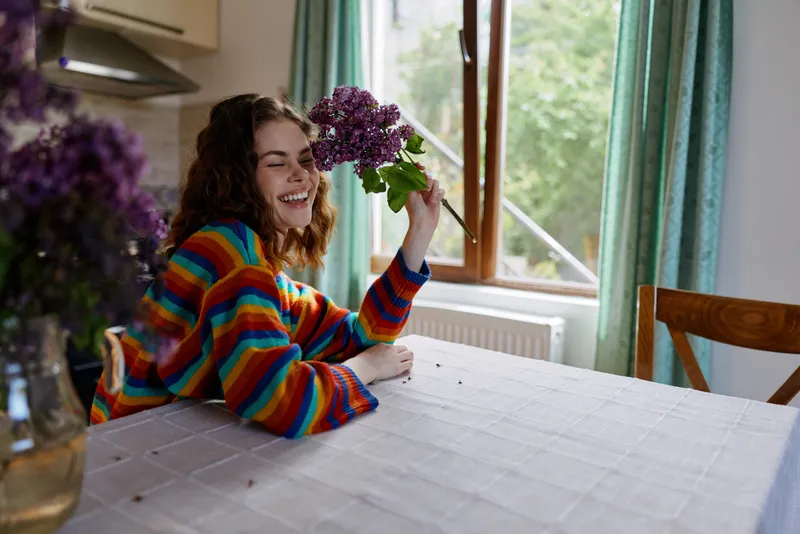 A woman sits at a sunlit kitchen table, holding a bouquet and smiling. A cozy home lifestyle moment highlighting warmth, color, and everyday joy in living spaces and daily routines.