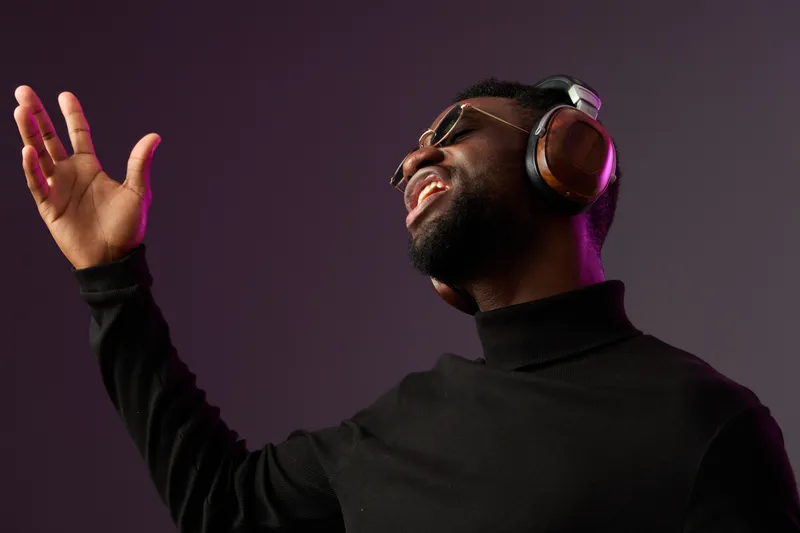 An African American man passionately singing with headphones on, wearing sunglasses, against a gradient background with purple and black tones