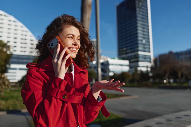 Young woman with curly hair wearing a red coat talks on a smartphone outdoors in an urban setting during sunny daytime with a joyful expression.