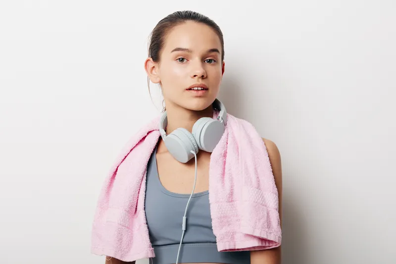 Woman in athletic wear with a pink towel around her neck and headphones, ready for a workout. Clean studio backdrop highlights fitness mood and focused performance.