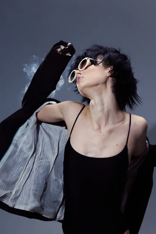 Woman wearing sunglasses and black dress poses dynamically with motion blur against dark background.