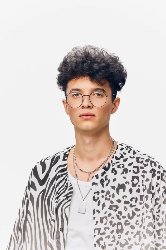 young man with curly hair and glasses wearing patterned shirt with abstract animal print on white background, casual style, confident expression
