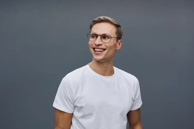 Smiling young man wearing glasses in casual white t-shirt looking away isolated on plain gray background studio portrait Portrait of a cheerful man Concept: happiness and confidence
