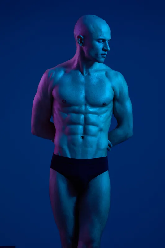 Athletic man posing in a dramatic light setting, showcasing his toned physique and strong presence. The image emphasizes fitness and strength with vibrant colors.