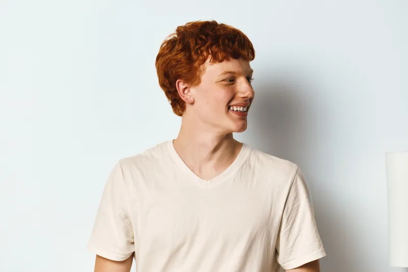 Young man with red hair smiling and looking to side wearing white t shirt in studio with plain background. Portrait of happy male adult with casual outfit and natural expression.