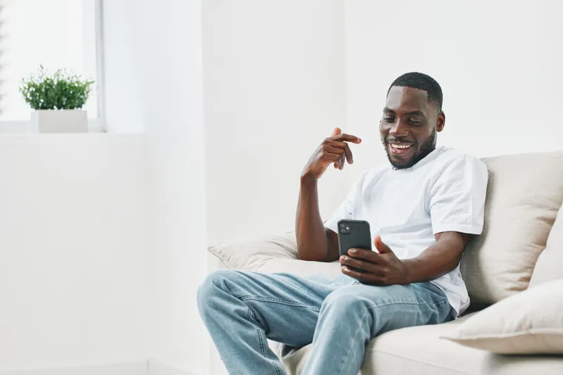 Happy young African man using smartphone in bright living room, casual outfit, smiling, enjoying leisure time, modern interior, relaxed atmosphere