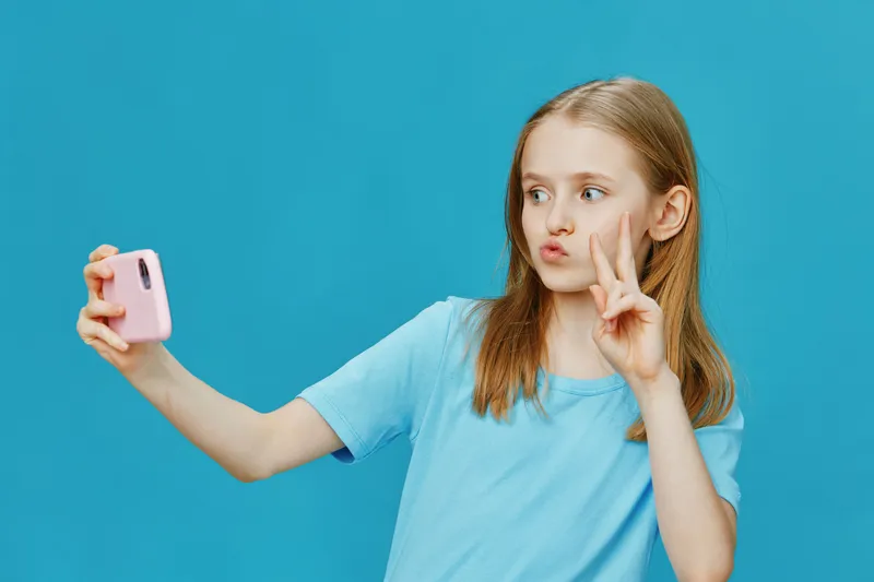 Young girl taking a selfie with a pink camera against a vibrant blue background, expressing joy and playfulness in casual attire, perfect for social media themes