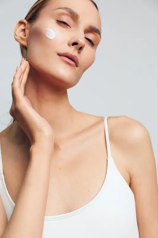 A young woman with clear skin applies cream to her face, showing skincare routine and beauty care in a bright studio setting with a soft neutral background.