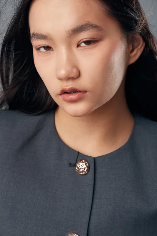 Portrait of young woman with clear skin and long dark hair wearing formal grey jacket with gold buttons in studio. Beauty, fashion and natural makeup with confident expression and soft lighting.