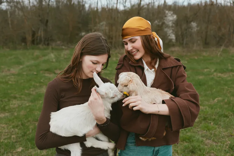 Two young women enjoy a lifestyle moment outdoors holding baby goats while smiling and connecting in casual autumn clothing on a cloudy day in a natural field setting.