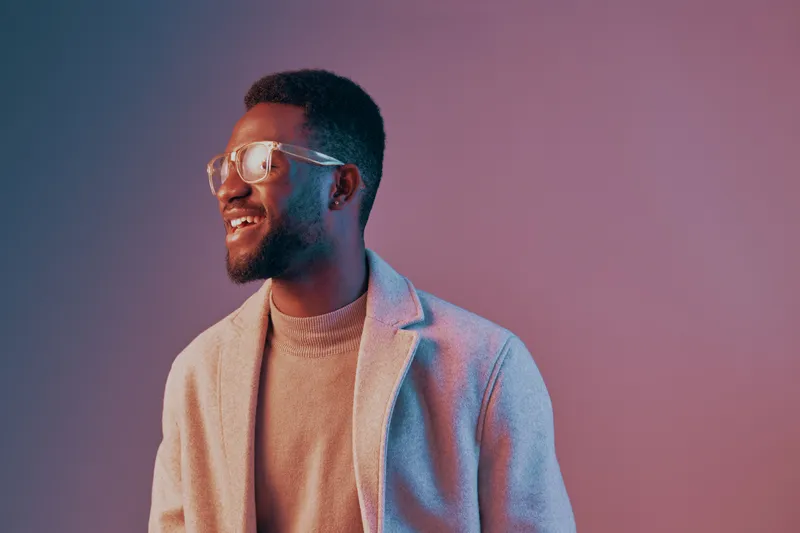Smiling man in glasses wearing a stylish gray coat and sweater, showcasing positivity and confidence against a colorful gradient background