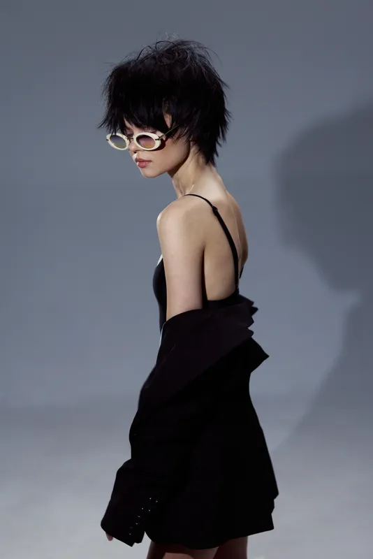 Young woman with short hair wearing sunglasses and black dress, stylish and modern pose on grey background.