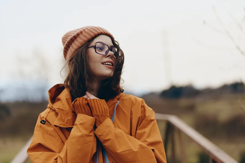 Woman lifestyle outdoors in autumn, smiling in orange jacket, knit hat, glasses, and scarf, warm sunshine and countryside landscape create a calm portrait concept with soft blur