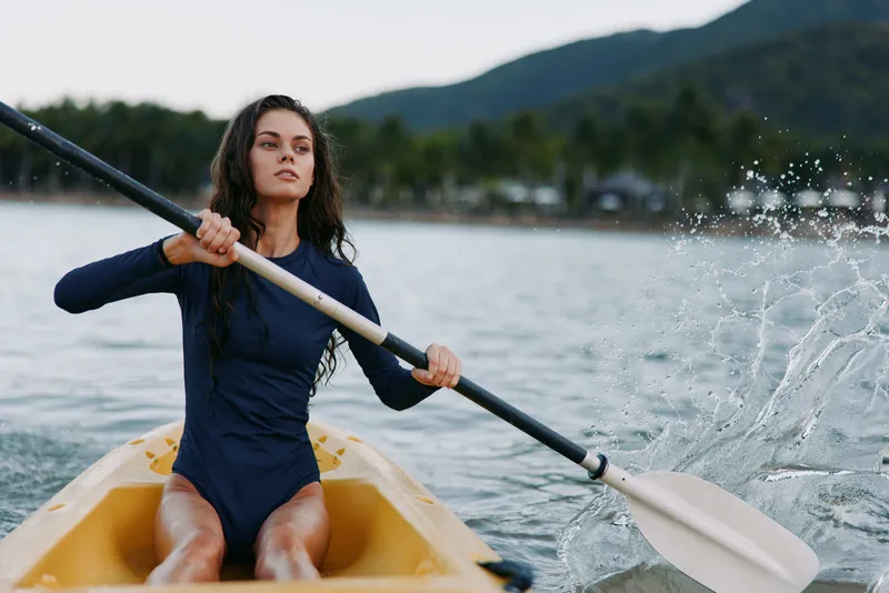A focused female rower sits in a yellow kayak, propelling with a long paddle as water splashes from the blade, highlighting strength, balance, and motion.