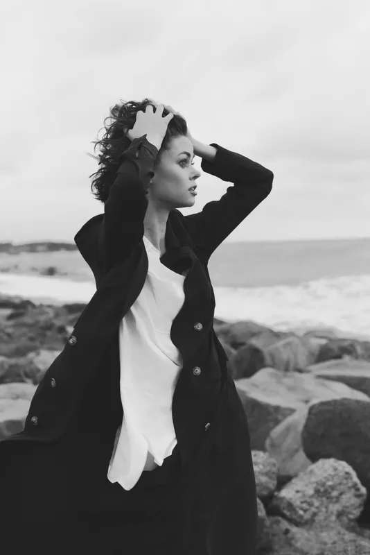 Wind swept seaside image shows a woman in a dark coat and white blouse, standing on rocks by the shore, hair in the wind, calm mood and cinematic monochrome tones