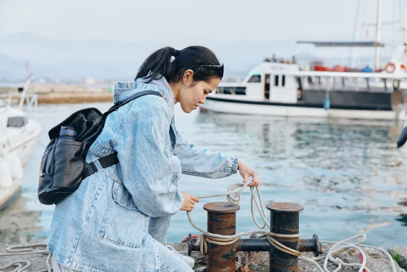 Woman with backpack handling rope at a marina dock, tying lines to bollards for mooring, outdoor scene with boats and calm water, casual denim jacket, focused and practical work