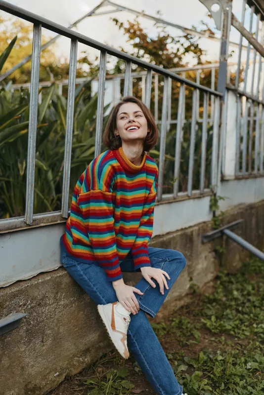 Young woman in a rainbow sweater and jeans sitting by a greenhouse railing, smiling with short hair in an outdoor lifestyle portrait, casual outfit and relaxed autumn mood.