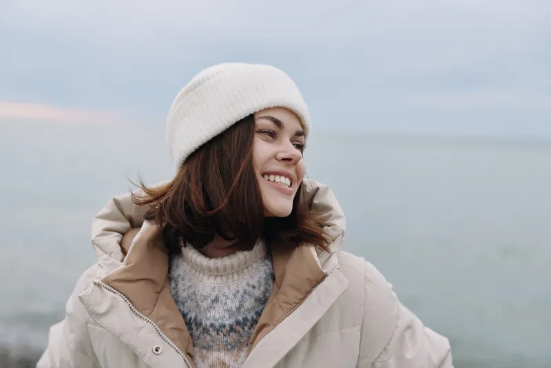 Woman in a warm winter coat and beanie smiles softly outdoors by the sea, capturing a calm coastal moment with a gentle breeze and pale sky.