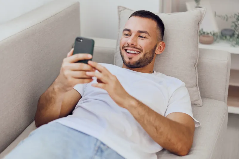 Smiling man enjoying a digital detox while casually using a smartphone, embodying a relaxed home environment and embracing self-care moments.
