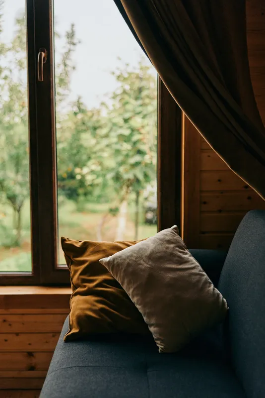 Cozy sofa with cushions by a large window, bathed in warm sunlight, a tranquil living space with wooden walls and soft fabrics creating an inviting atmosphere.