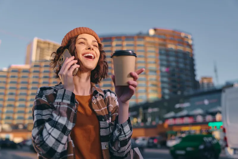 woman coffee phone city smile beanie walking morning urban candid lifestyle authenticity golden hour glow mindful living emotional storytelling joyous young woman laughing on phone holding takeaway