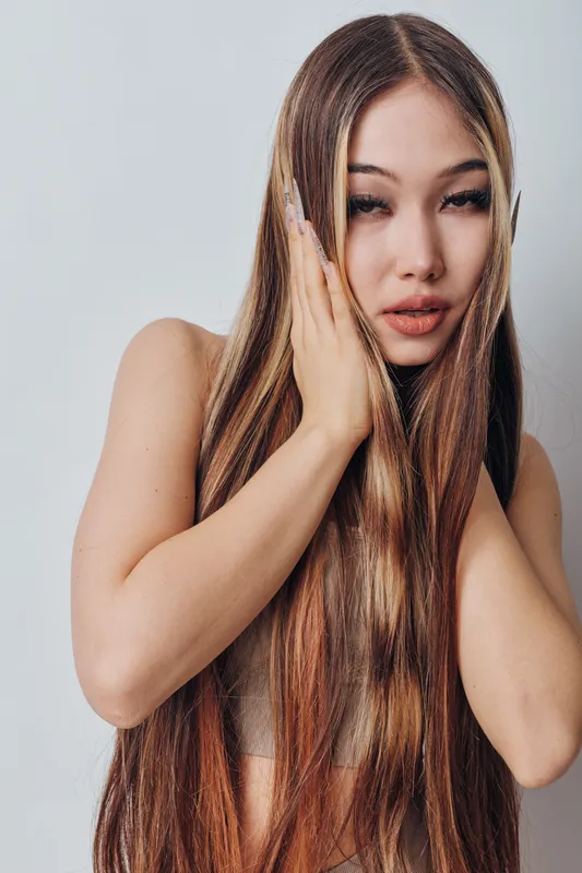Young woman with long straight hair touching face with hands in studio. Beauty portrait of female model with healthy hair and natural makeup on light background.