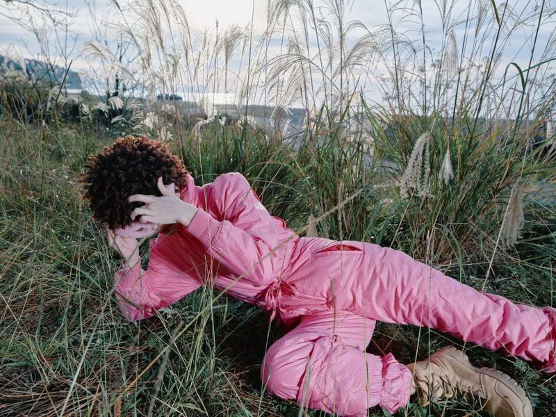 Outdoor scene with a person in a pink jumpsuit lounging in tall grass, relaxed pose, natural light, casual fashion vibe in a nature setting, conveying freedom and outdoor style.